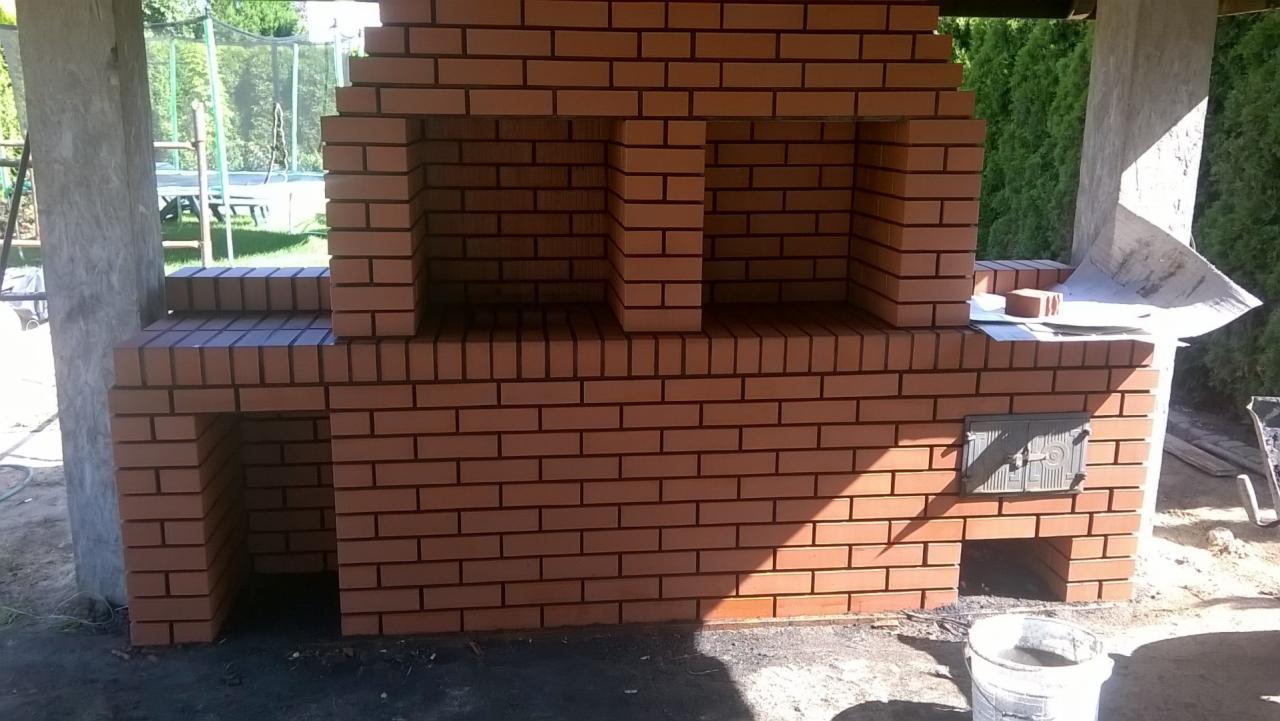 Nowo wymurowany, ceglany grill ogrodowy z wędzarnią, z widoczną metalową klapą i otworami wentylacyjnymi, usytuowany pod zadaszeniem z betonowym słupem, w tle zieleń ogrodu.