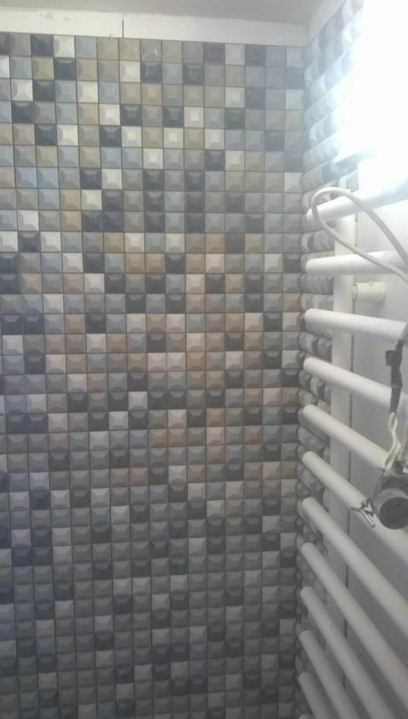 Ściana wyłożona mozaiką z trójwymiarowych, kwadratowych płytek w odcieniach szarości, beżu i błękitu, z grzejnikiem drabinkowym po prawej stronie, widoczne jasne światło z okna.