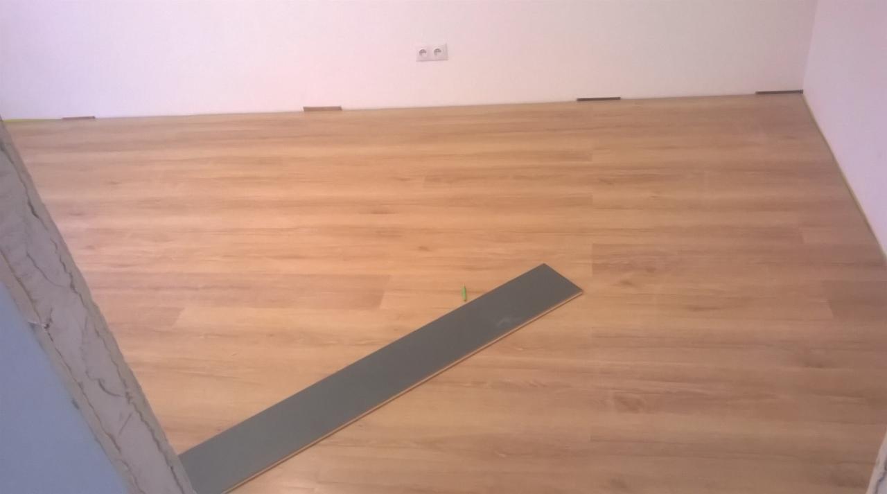 Układanie paneli podłogowych: widok na fragment ułożonej podłogi z paneli imitujących drewno, pojedynczy panel leżący na podłodze, widoczne dystanse przy ścianie, gniazdko elektryczne na ścianie...