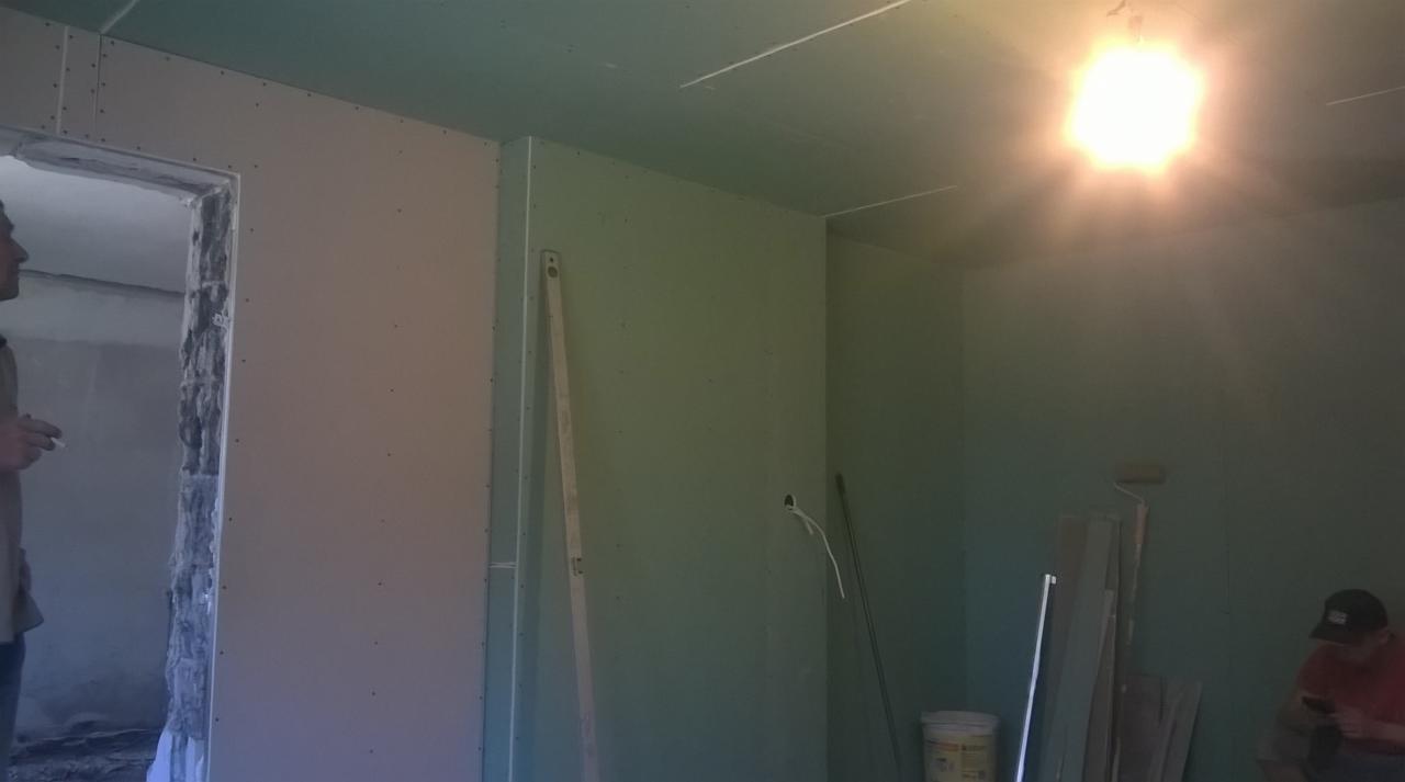 Wnętrze w trakcie wykańczania, ściany pokryte płytami gipsowo-kartonowymi, widoczne narzędzia i materiały budowlane, fragment otworu drzwiowego w surowym stanie, pracownik w tle.