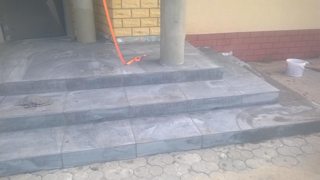 Wykończone, szare, ceramiczne schody wejściowe do budynku z kolumnami, widoczny fragment elewacji z cegły i wiadro po farbie.