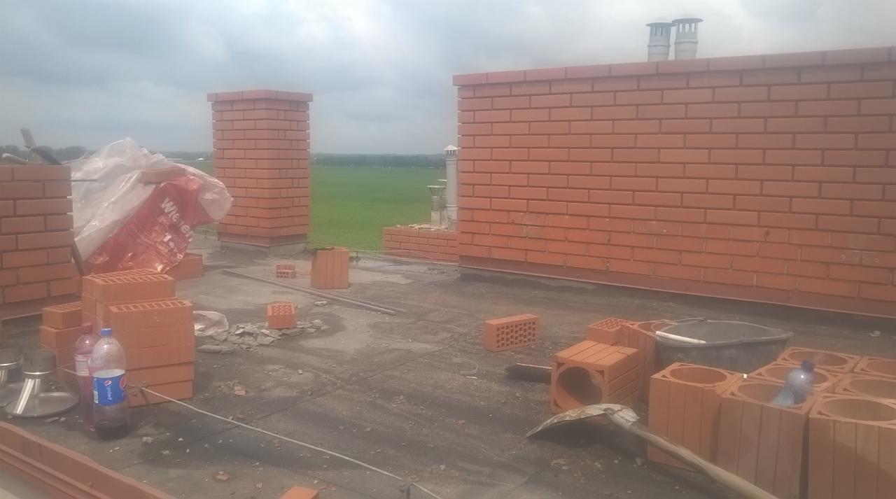 Budowa komina i ściany z czerwonej cegły na dachu, widoczne materiały budowlane, narzędzia i butelki z wodą, w tle zielone pole.