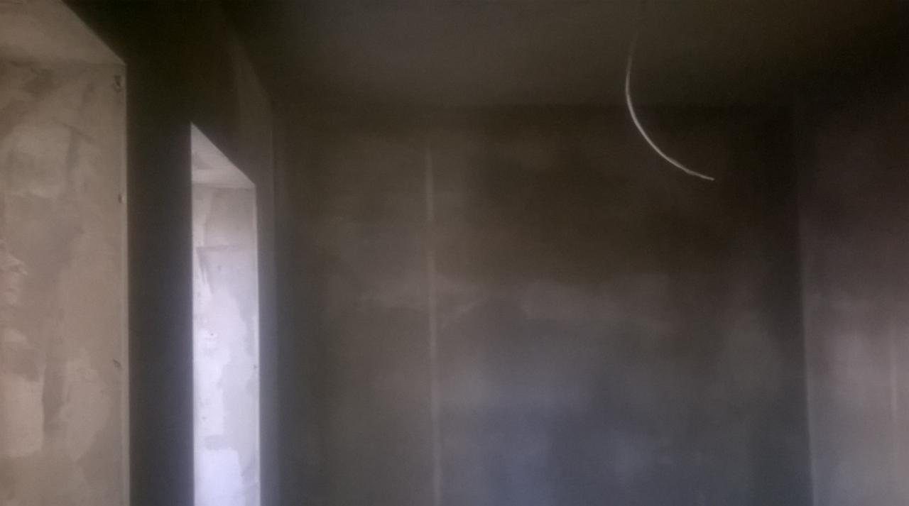 Świeżo nałożony szary tynk na ścianach i suficie w pomieszczeniu w trakcie remontu. Widoczny kabel zwisający z sufitu i fragment otworu drzwiowego.