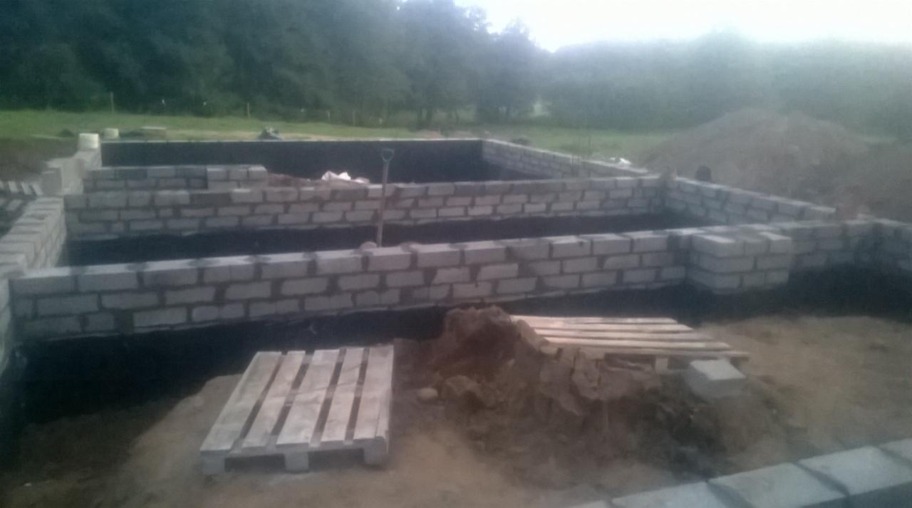 Wykop pod fundamenty z częściowo wymurowanymi ścianami z bloczków betonowych, widoczne palety i łopata w wykopie, teren budowy w otoczeniu zieleni.