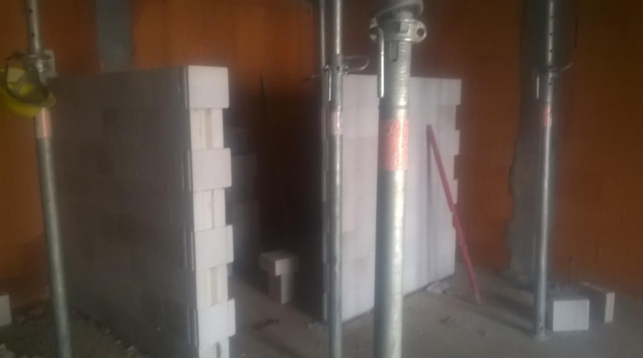 Wznoszenie ściany z szarych bloczków z białymi wstawkami, podpartej metalowymi stemplami budowlanymi w surowym wnętrzu budynku.