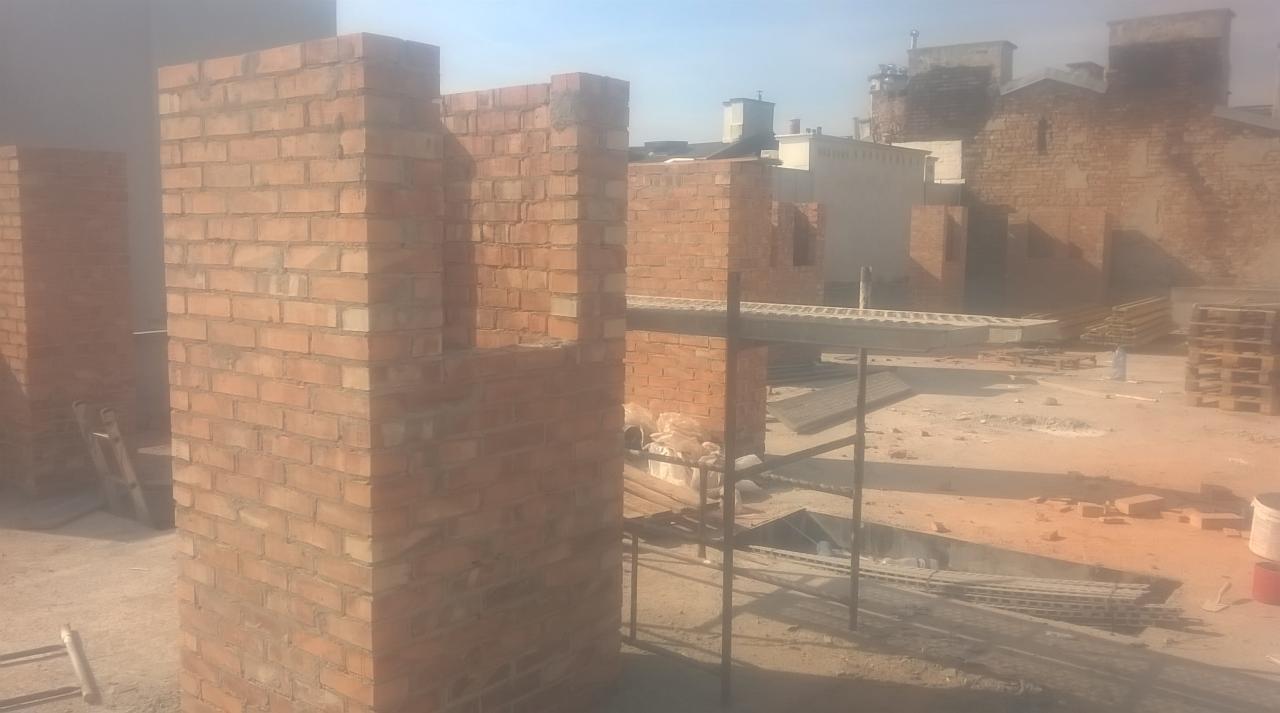 Wznoszone z czerwonej cegły ściany na budowie, widoczne rusztowania i materiały budowlane w tle.