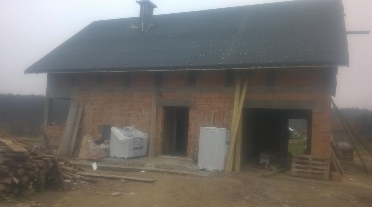 Surowy budynek z czerwonej cegły z ciemnym dachem, widoczne pakiety materiałów budowlanych, drewno i nieotynkowane ściany.