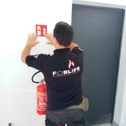 Forlife Bezpieczeństwo Pożarowe S.C. - Mężczyzna w czarnej koszulce z logo firmy FORLIFE montuje znak przeciwpożarowy na białej ścianie, widoczny gaśnica i poziomica laserowa.