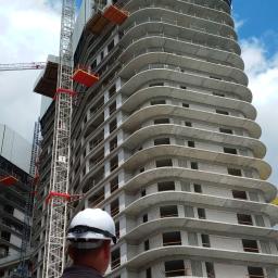 Forlife Bezpieczeństwo Pożarowe S.C. - Pracownik w kasku na budowie, widok na wysoki budynek w trakcie budowy z żurawiem, logo firmy FORLIFE na ubraniu.