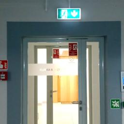 Forlife Bezpieczeństwo Pożarowe S.C. - Oznakowane wyjście ewakuacyjne z zielonym znakiem nad drzwiami, czerwony przycisk alarmowy i instrukcja wzywania pomocy na ścianie, klawiatura numeryczna dostępu.