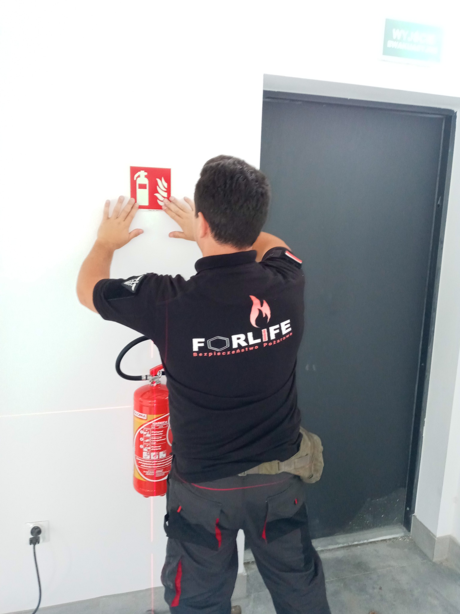 Mężczyzna w czarnej koszulce z logo firmy FORLIFE montuje znak przeciwpożarowy na białej ścianie, widoczny gaśnica i poziomica laserowa.