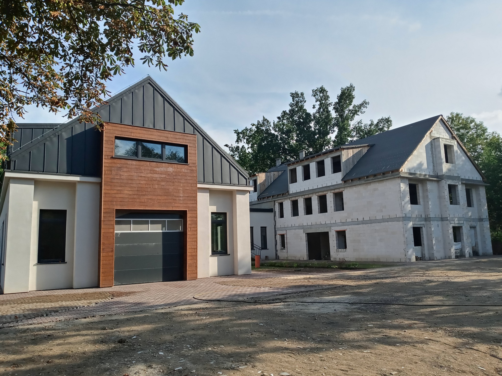 Dwa budynki: jeden ukończony z szarą bramą garażową i drewnianą elewacją, drugi w stanie surowym zamkniętym, z widocznymi pustakami i oknami bez szyb.
