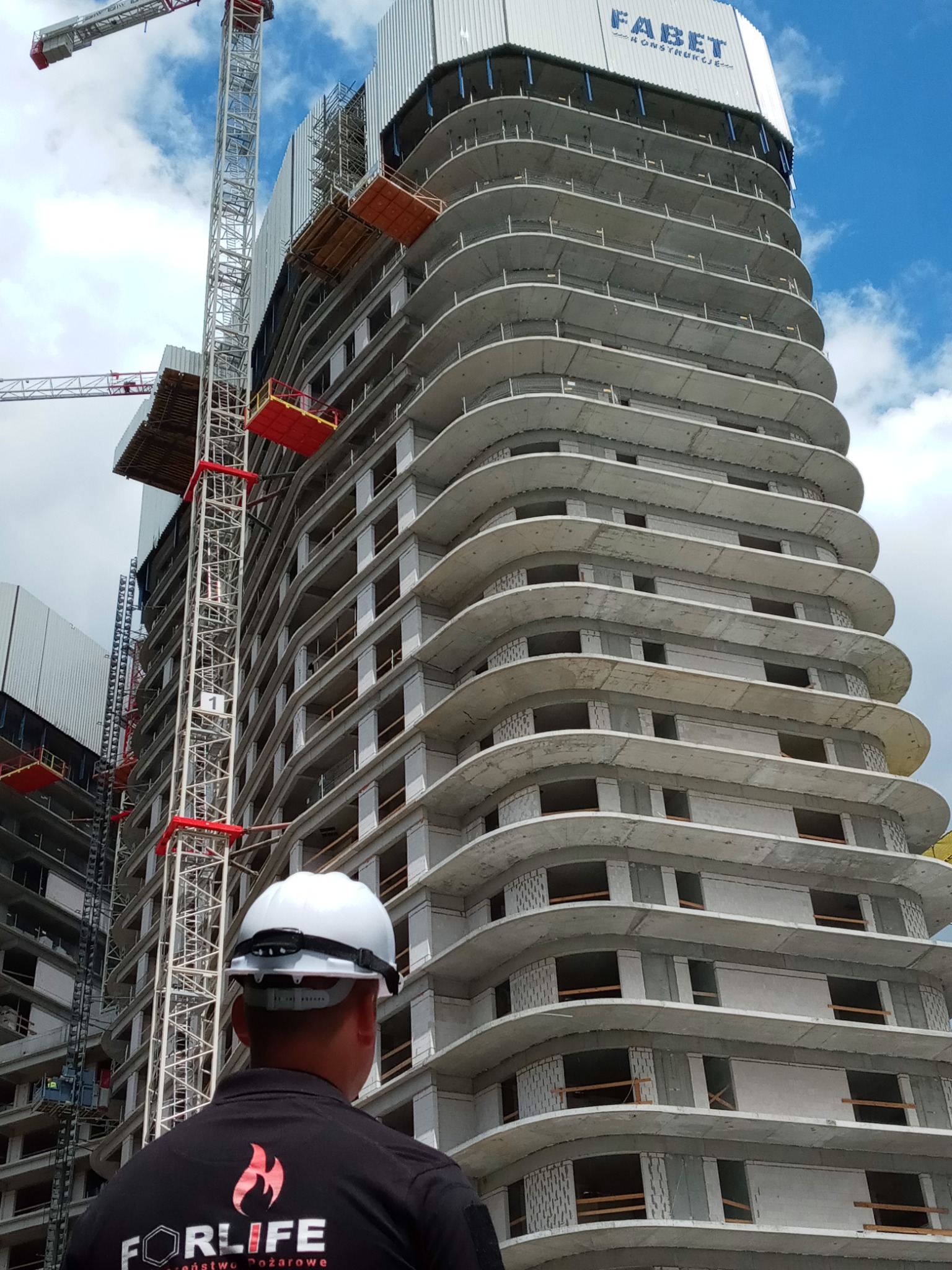 Pracownik w kasku na budowie, widok na wysoki budynek w trakcie budowy z żurawiem, logo firmy FORLIFE na ubraniu.