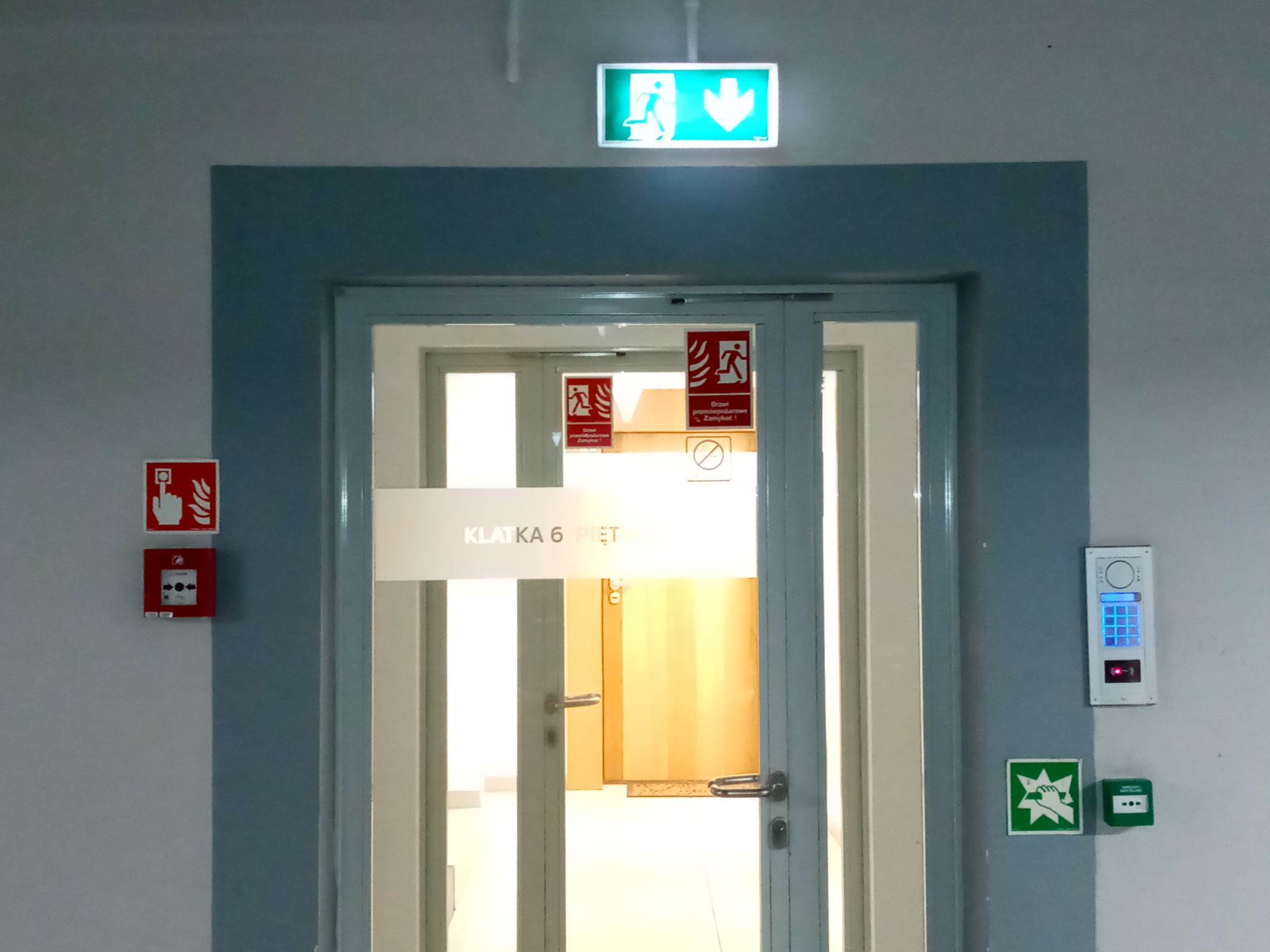Oznakowane wyjście ewakuacyjne z zielonym znakiem nad drzwiami, czerwony przycisk alarmowy i instrukcja wzywania pomocy na ścianie, klawiatura numeryczna dostępu.