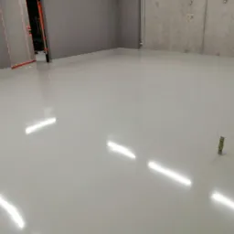 Jasnoszara, błyszcząca posadzka w pomieszczeniu z szarą ścianą i betonową ścianą w tle. Na posadzce widoczne odbicia światła i mały, pionowy element owinięty drutem.