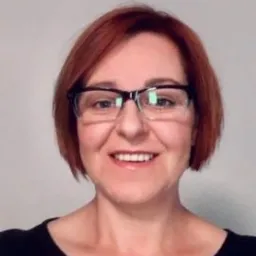 Uśmiechnięta kobieta w okularach, z krótkimi rudymi włosami, na neutralnym tle. Portret biznesowy w czarnej bluzce.