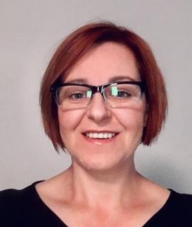 Uśmiechnięta kobieta w okularach, z krótkimi rudymi włosami, na neutralnym tle. Portret biznesowy w czarnej bluzce.