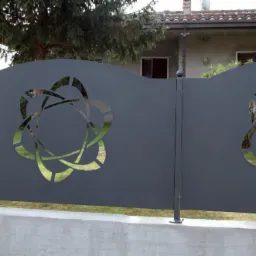 Szary metalowy panel ogrodzeniowy z wyciętym abstrakcyjnym wzorem na tle domu i zieleni.