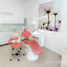 Jasne wnętrze gabinetu z fotelem w odcieniach różu, białymi szafkami i sprzętem medycznym, na ścianie obraz z motywem kwiatowym.