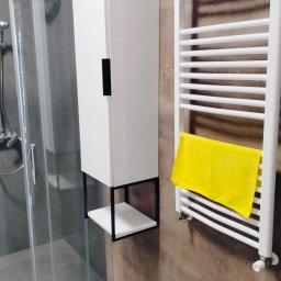 MAZUR usługi remontowo-budowlane Andrzej Walczak - Nowoczesna łazienka z kabiną prysznicową, białą szafką wiszącą z czarnymi elementami, grzejnikiem drabinkowym z powieszonym żółtym ręcznikiem i szarą płytką imitującą beton na ścianach.