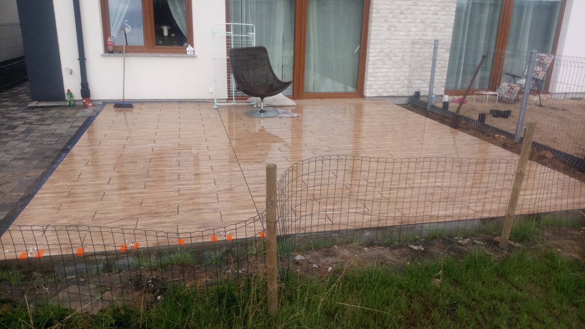 Świeżo ułożona, brązowa, imitująca drewno, płytka tarasowa na zewnątrz domu z widocznymi plastikowymi elementami poziomującymi przy krawędziach; w tle krzesło i suszarka na pranie.