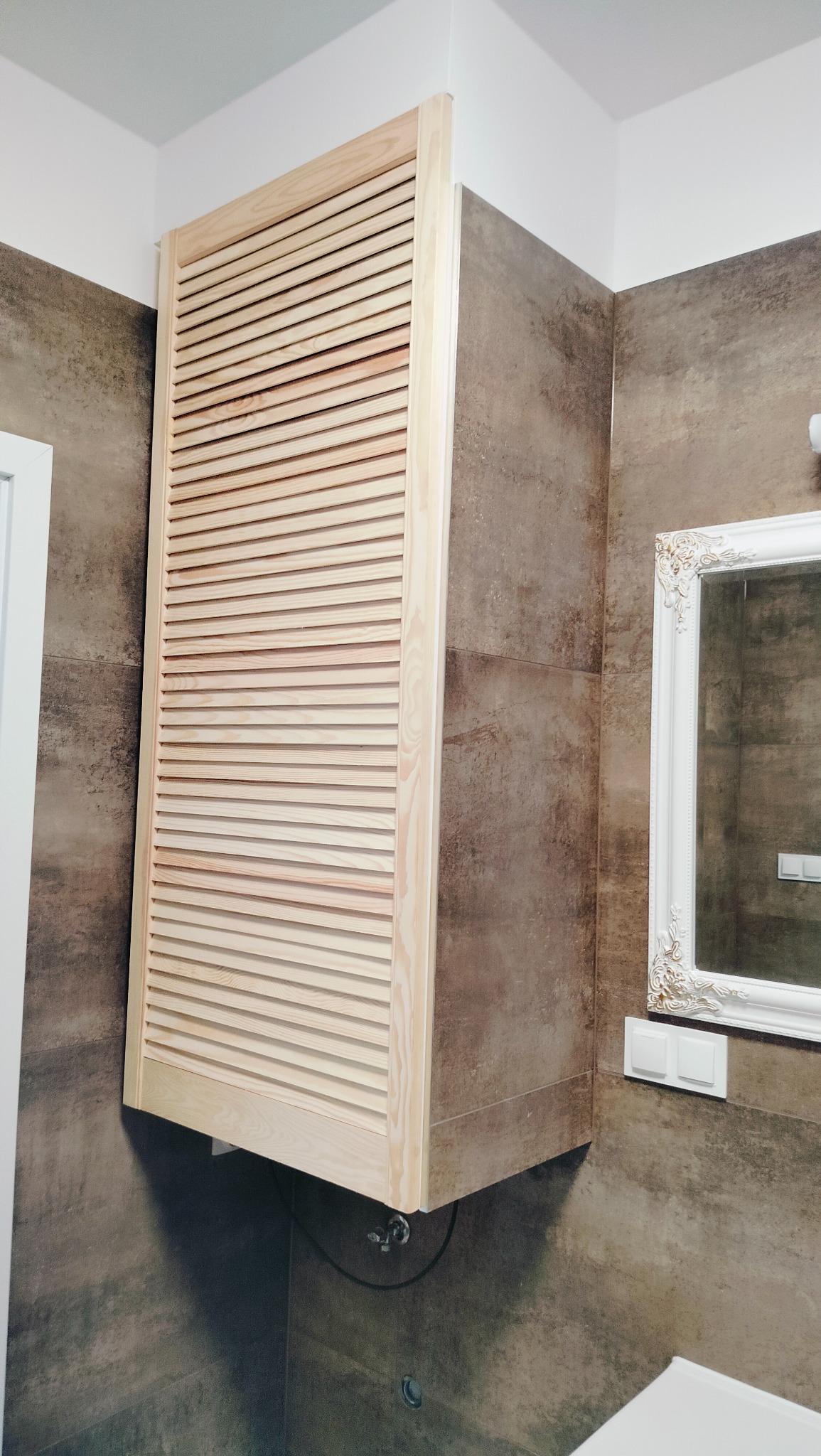 Drewniana szafka z żaluzjowymi drzwiczkami, zamontowana w rogu łazienki, na tle płytek w odcieniach brązu, obok lustro w ozdobnej, białej ramie.