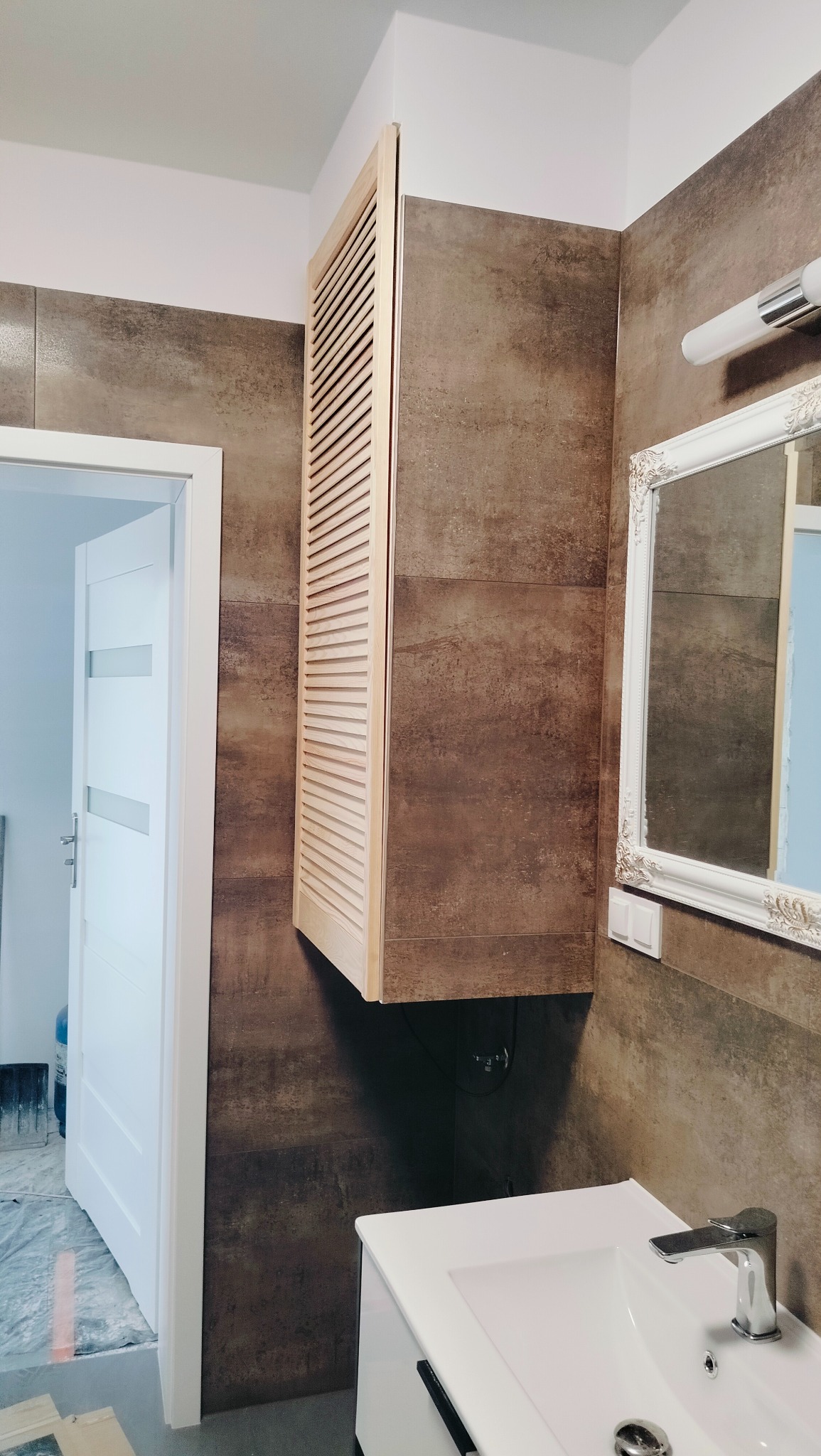 Nowoczesna łazienka w Szczecinie z brązowymi płytkami imitującymi kamień, białą umywalką z szafką, lustrem w ozdobnej ramie oraz drewnianą szafką z żaluzjowymi drzwiczkami.