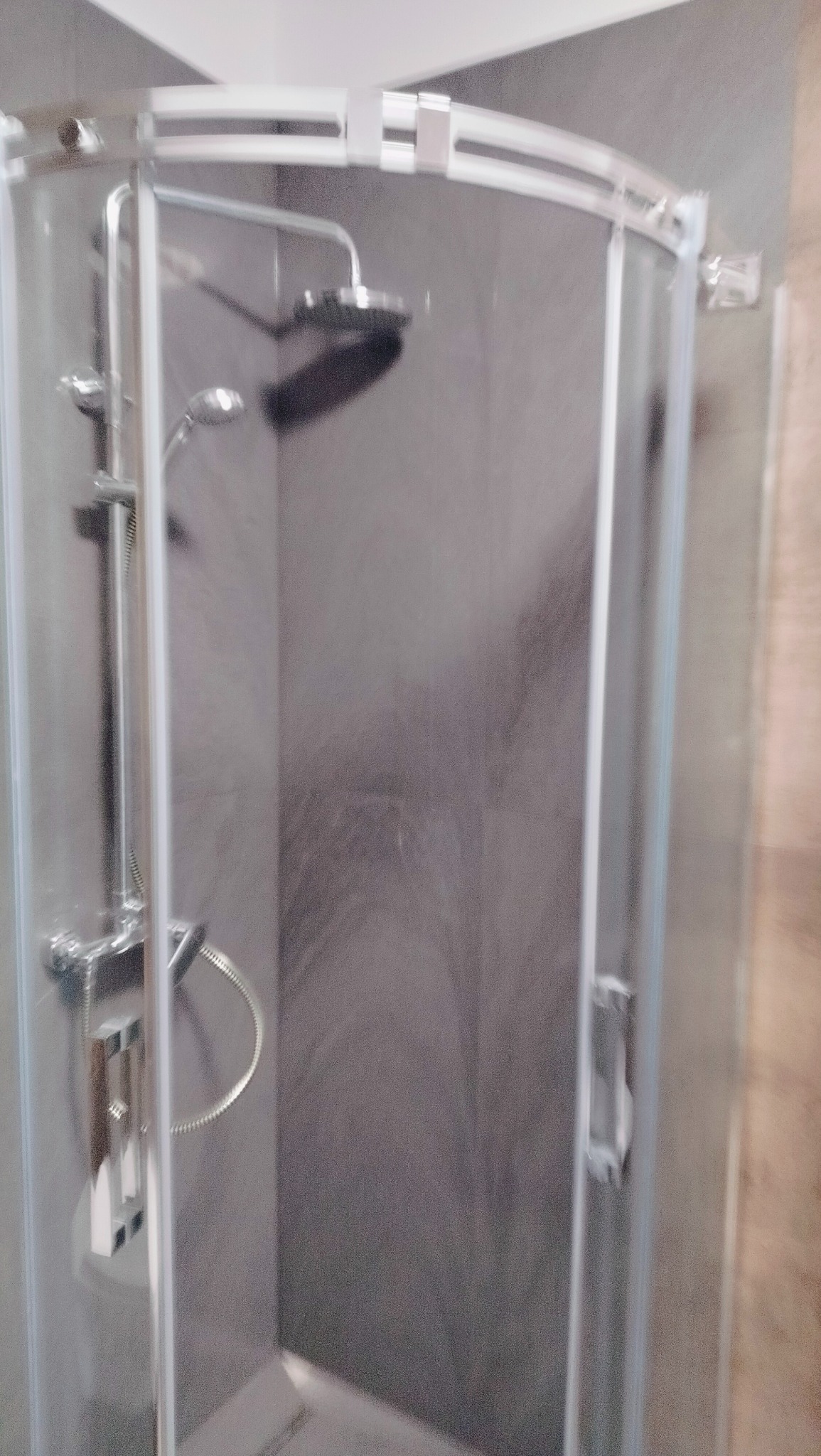 Półokrągła kabina prysznicowa z chromowaną armaturą, deszczownicą i szarą, teksturowaną ścianą w tle.