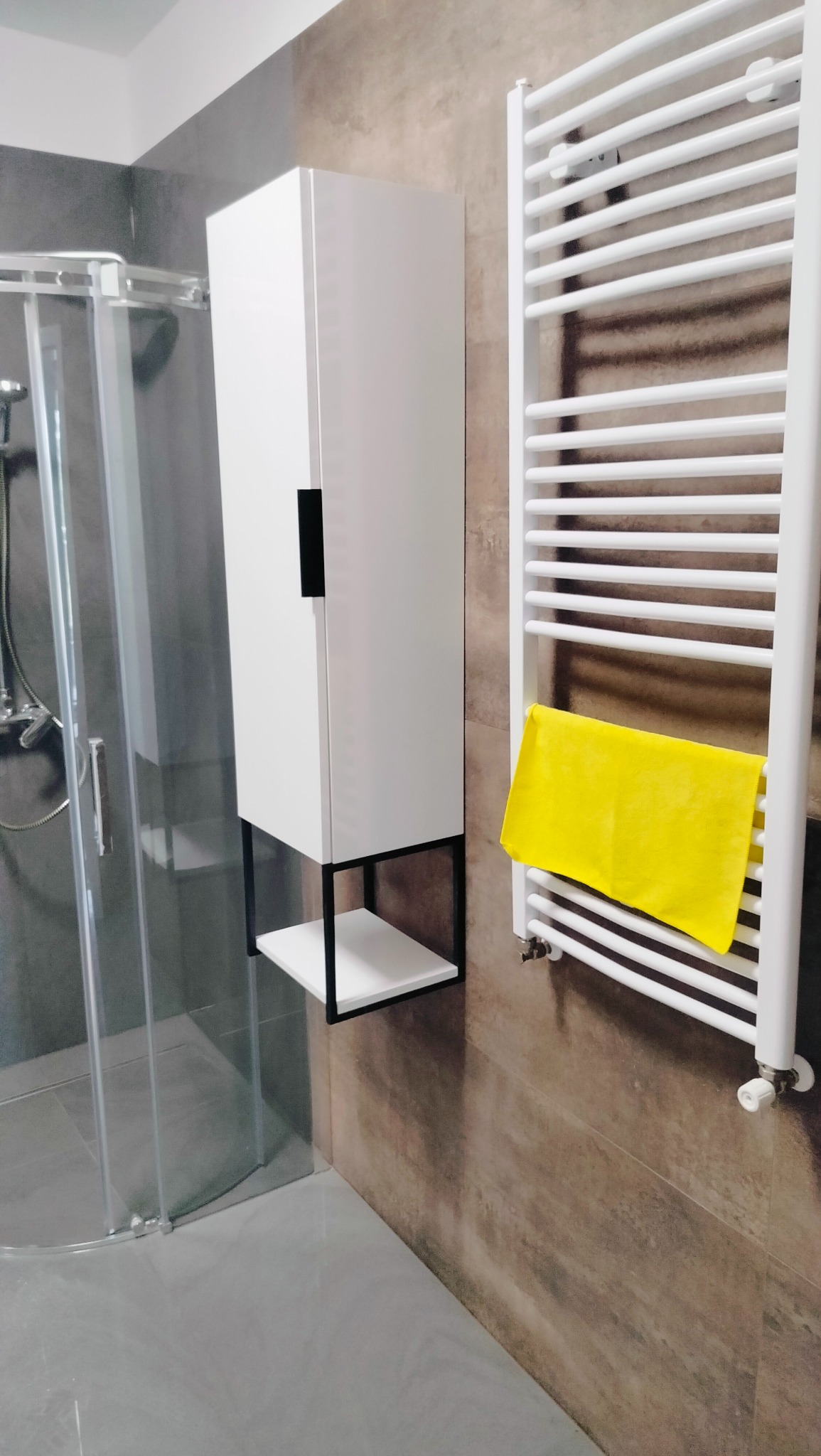 Nowoczesna łazienka z kabiną prysznicową, białą szafką wiszącą z czarnymi elementami, grzejnikiem drabinkowym z powieszonym żółtym ręcznikiem i szarą płytką imitującą beton na ścianach.