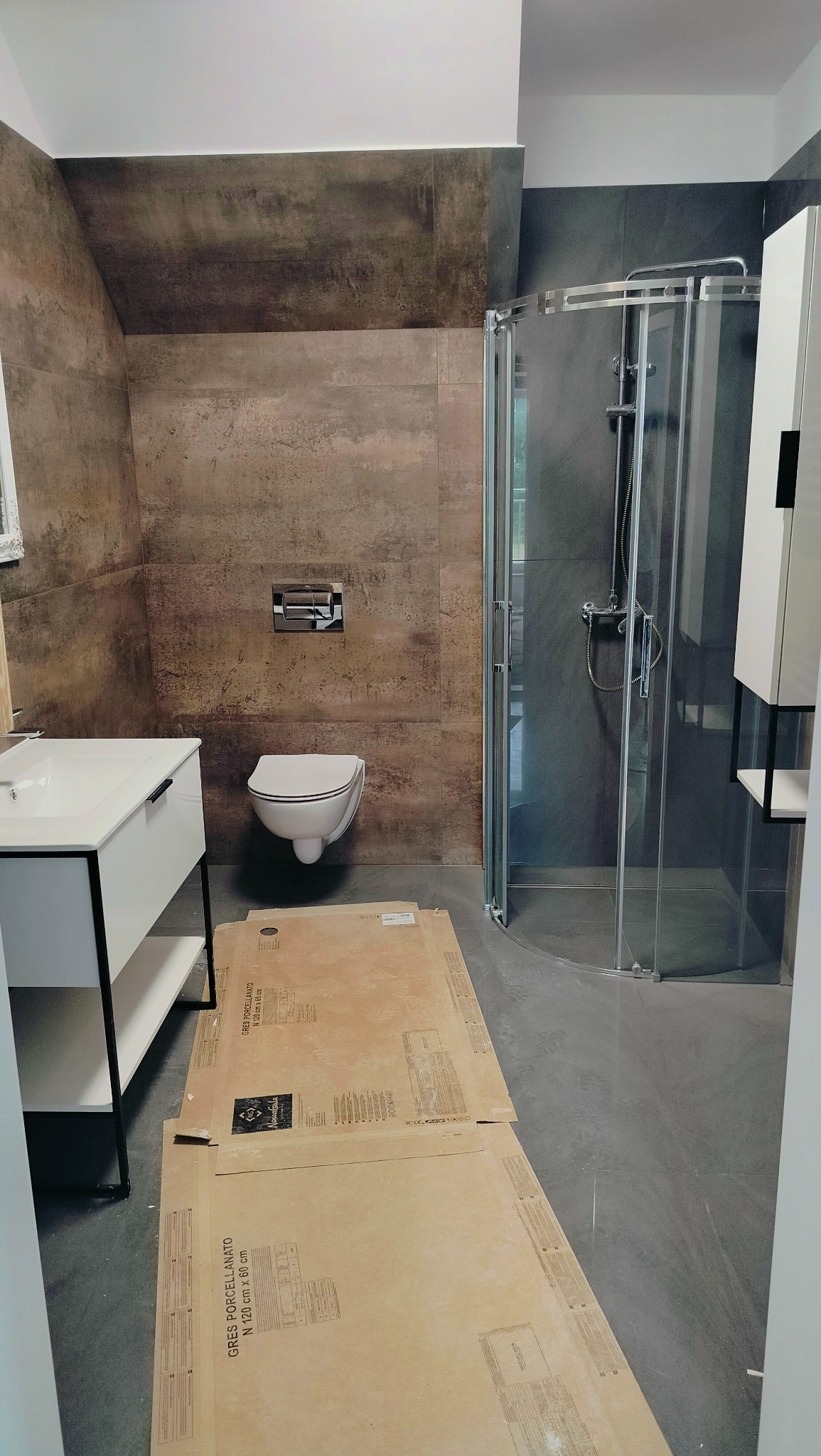 Nowoczesna łazienka w trakcie wykańczania, widoczna kabina prysznicowa, podwieszana toaleta, umywalka z szafką oraz zabezpieczone kartonami płytki podłogowe przed dalszymi pracami.