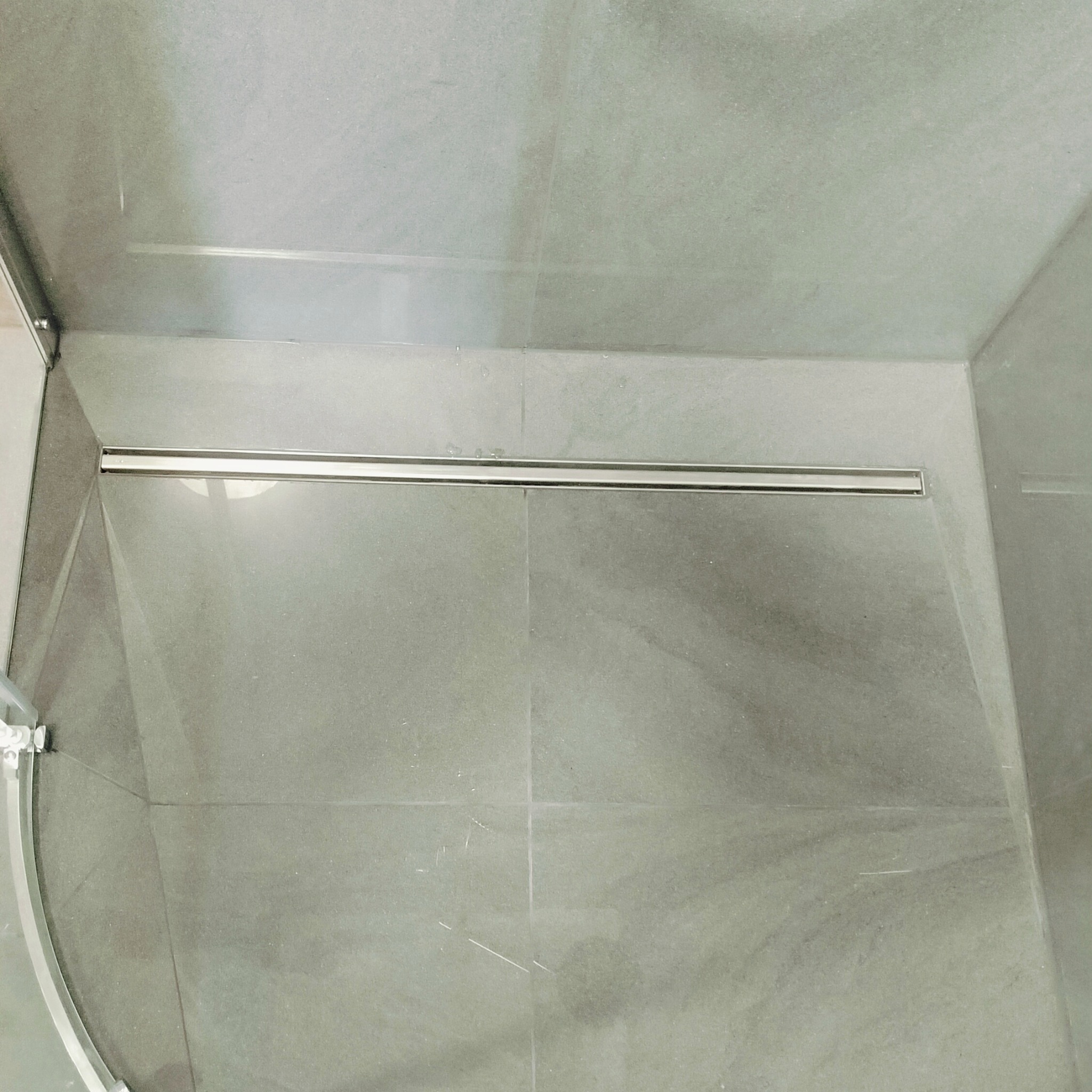 Szare płytki w kabinie prysznicowej z odpływem liniowym w podłodze, widoczna szklana ściana kabiny.