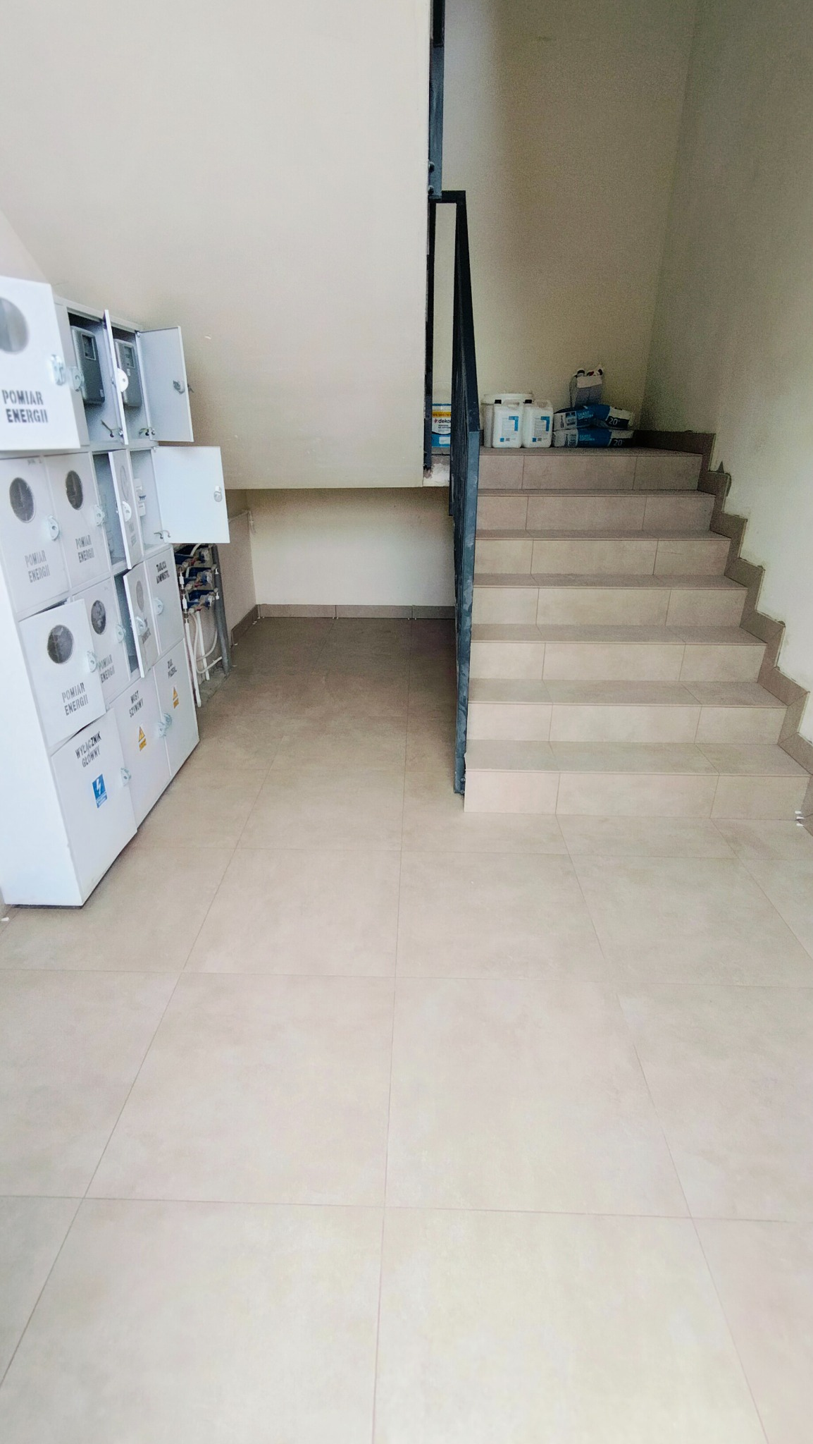 Klatka schodowa z beżowymi płytkami podłogowymi i schodami, białe szafki z licznikami energii po lewej stronie, farby i materiały budowlane na schodach w tle.