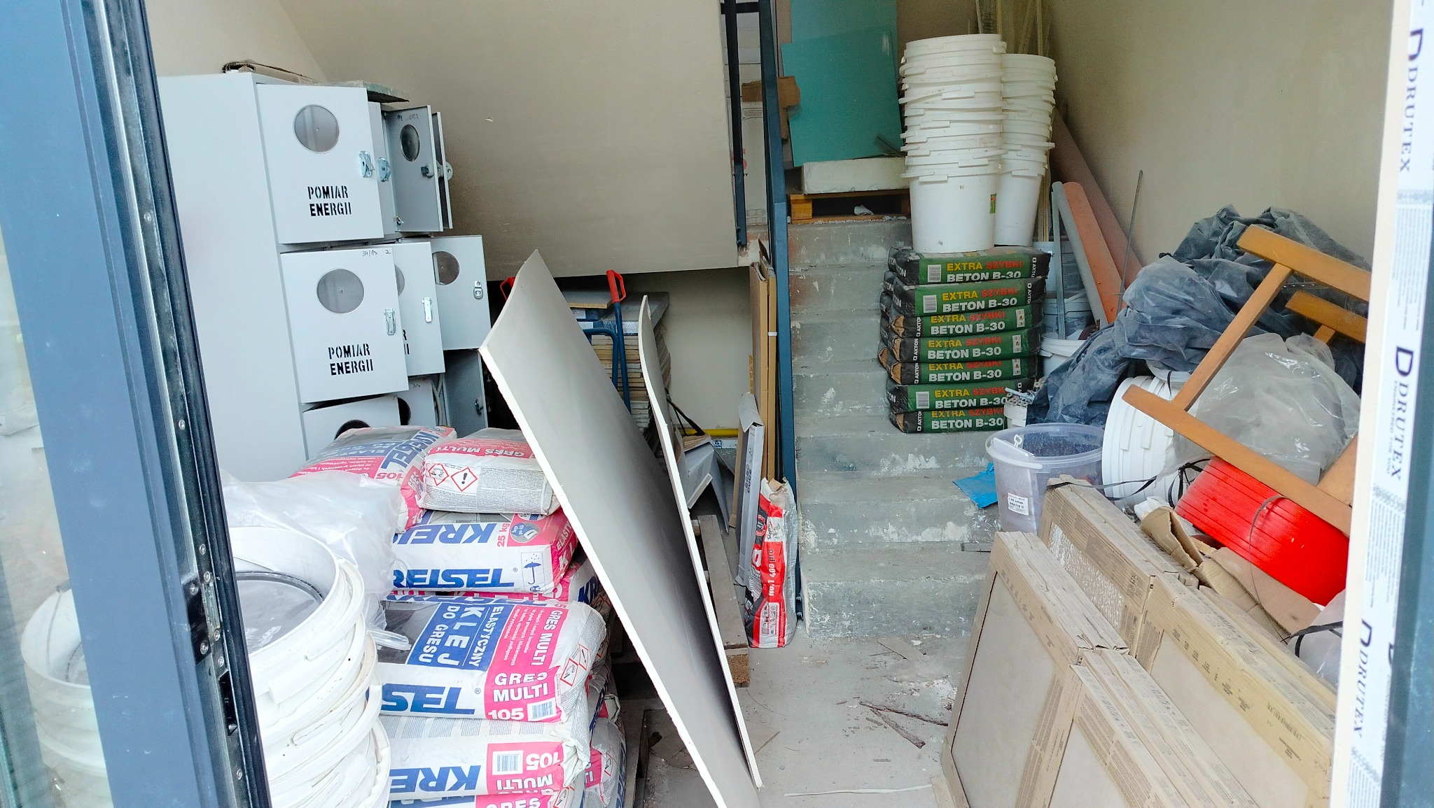 Sterta materiałów budowlanych w pomieszczeniu: worki z klejem, płyty karton gips, wiadra, worki z betonem, skrzynki z napisem 'Pomiar Energii' oraz fragment schodów.