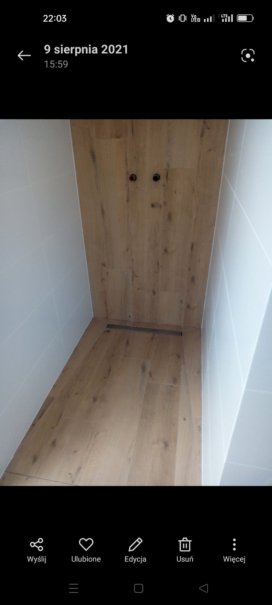 Nowoczesna kabina prysznicowa walk-in z drewnopodobnymi płytkami na ścianie i podłodze, odpływ liniowy w podłodze, białe płytki na ścianach bocznych i widoczne przyłącza wodne.