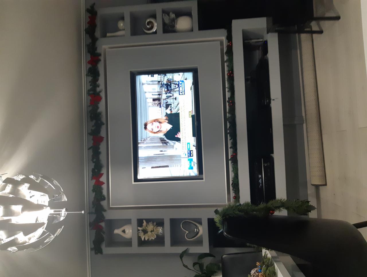 Telewizor wbudowany w szarą zabudowę meblową z półkami dekoracyjnymi, ozdobiony świąteczną girlandą z czerwonymi kokardkami, oświetlony nowoczesną lampą wiszącą.