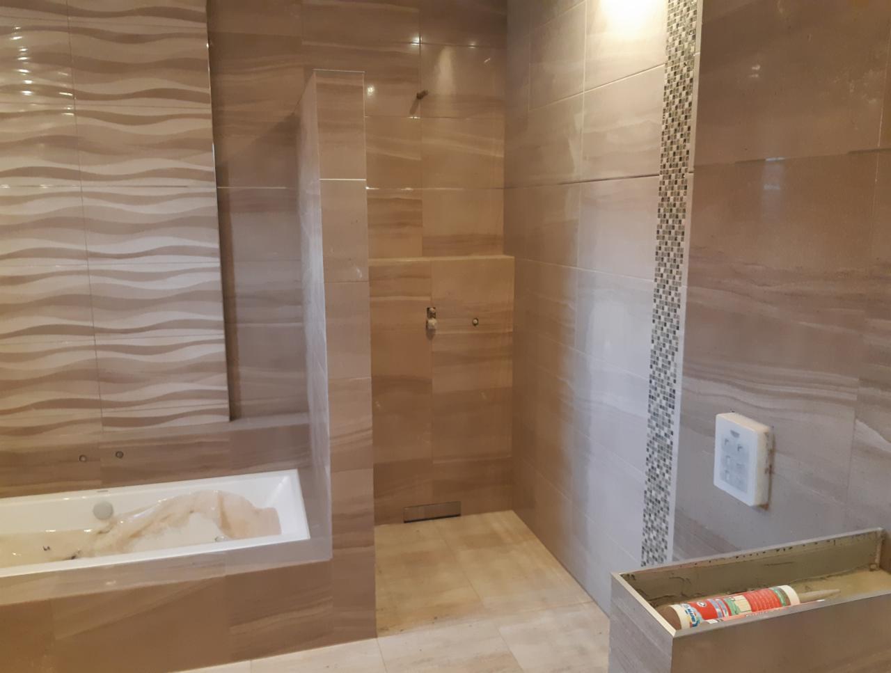 Nowoczesna łazienka w trakcie wykańczania, z beżowymi płytkami imitującymi marmur, widoczna wanna zabezpieczona folią oraz fragment brodzika z odpływem liniowym, w tle dekoracyjny pas mozaiki.