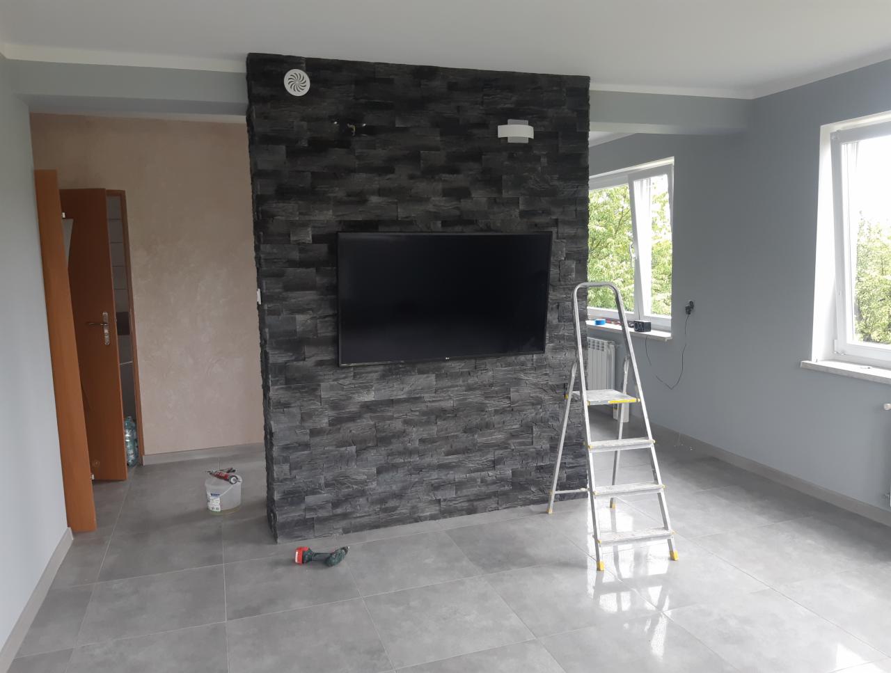 Pokój po remoncie z szarą ścianą z kamienia dekoracyjnego, na której wisi telewizor, szare ściany, podłoga z szarych płytek, drabina i narzędzia malarskie.