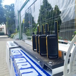 BEST HOME MARIUSZ MIKOŁAJCZYK - Cztery radiotelefony Motorola stojące w rzędzie na palecie materiałów budowlanych, widoczne ogrodzenie budowlane w tle.