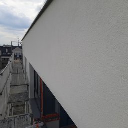 BEST HOME MARIUSZ MIKOŁAJCZYK - Świeżo otynkowana, jasna elewacja budynku wielorodzinnego, widoczna z poziomu rusztowania z siatką zabezpieczającą, na tle zabudowy miejskiej i pochmurnego nieba.
