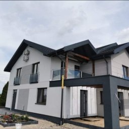 BEST HOME MARIUSZ MIKOŁAJCZYK - Dwupiętrowy dom z jasnoszarą elewacją, ciemnoszarą stolarką okienną i dachem, balkonami z szklanymi balustradami i zadaszeniem tarasu w kolorze grafitowym, widoczne dwa klimatyzatory na ścianie...