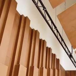 BEST HOME MARIUSZ MIKOŁAJCZYK - Akustyczna ściana z drewnianych paneli o nieregularnej wysokości, oświetlenie sceniczne na czarnej konstrukcji kratownicowej pod sufitem w sali widowiskowej.