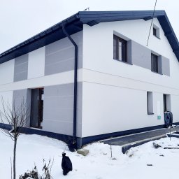BEST HOME MARIUSZ MIKOŁAJCZYK - Dwukondygnacyjny dom z białą elewacją i szarymi akcentami w zimowej scenerii, widoczne nowe rynny i fragment chodnika z kostki, małe drzewko na pierwszym planie.