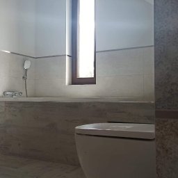 BEST HOME MARIUSZ MIKOŁAJCZYK - Jasna łazienka na poddaszu z oknem w drewnianej ramie, wykończona płytkami imitującymi marmur, z widoczną toaletą i wanną z prysznicem.