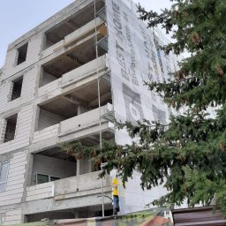 BEST HOME MARIUSZ MIKOŁAJCZYK - Surowy budynek wielopiętrowy w trakcie budowy, częściowo zasłonięty siatką z reklamą firmy budowlanej, widok z dołu, na pierwszym planie gałęzie drzewa iglastego.