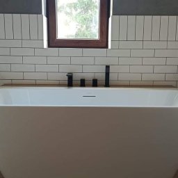 BEST HOME MARIUSZ MIKOŁAJCZYK - Nowoczesna biała wanna wolnostojąca z czarną armaturą na tle ściany wyłożonej białymi, prostokątnymi płytkami, drewniany blat pod oknem.
