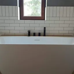 Nowoczesna biała wanna wolnostojąca z czarną armaturą na tle ściany wyłożonej białymi, prostokątnymi płytkami, drewniany blat pod oknem.