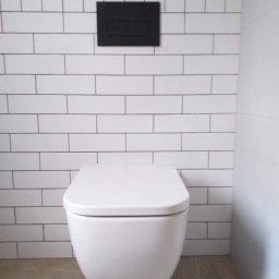 BEST HOME MARIUSZ MIKOŁAJCZYK - Nowoczesna toaleta podwieszana z czarnym przyciskiem spłukującym na ścianie wyłożonej białymi, prostokątnymi płytkami z ciemną fugą i podłogą imitującą drewno.