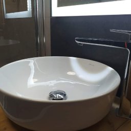 BEST HOME MARIUSZ MIKOŁAJCZYK - Okrągła, biała umywalka nablatowa z chromowaną baterią o minimalistycznym designie, ustawiona na blacie z jasnego drewna, z widoczną kabiną prysznicową w tle.