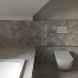 BEST HOME MARIUSZ MIKOŁAJCZYK - Wykończona łazienka na poddaszu z beżowymi płytkami imitującymi kamień, podwieszana toaleta z białym przyciskiem spłukującym i zabudowana wanna z marmurowym blatem.
