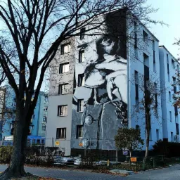 Wielkoformatowy portret na ścianie budynku mieszkalnego, częściowo pokryty tekstem, widoczny z poziomu ulicy z drzewem na pierwszym planie.